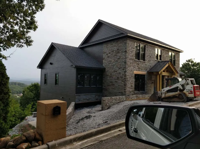 New house under construction with stone facade, dark roof, and a small porch; a tractor is parked nearby.
