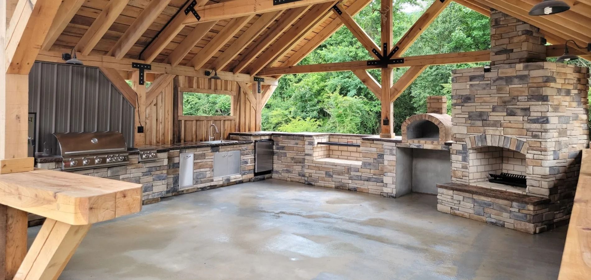 Outdoor kitchen with stone accents, a built-in grill, and a pizza oven under a wood-beamed roof.