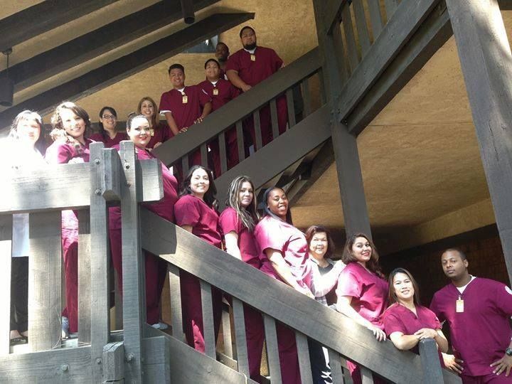 Group of healthcare workers in maroon scrubs pose on wooden stairs.