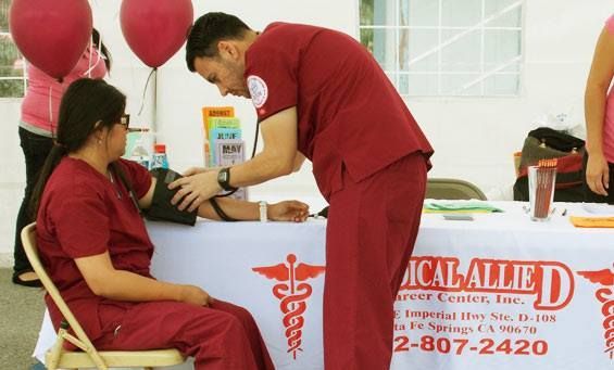A medical professional in red scrubs checks a patient's blood pressure outdoors at a health fair.