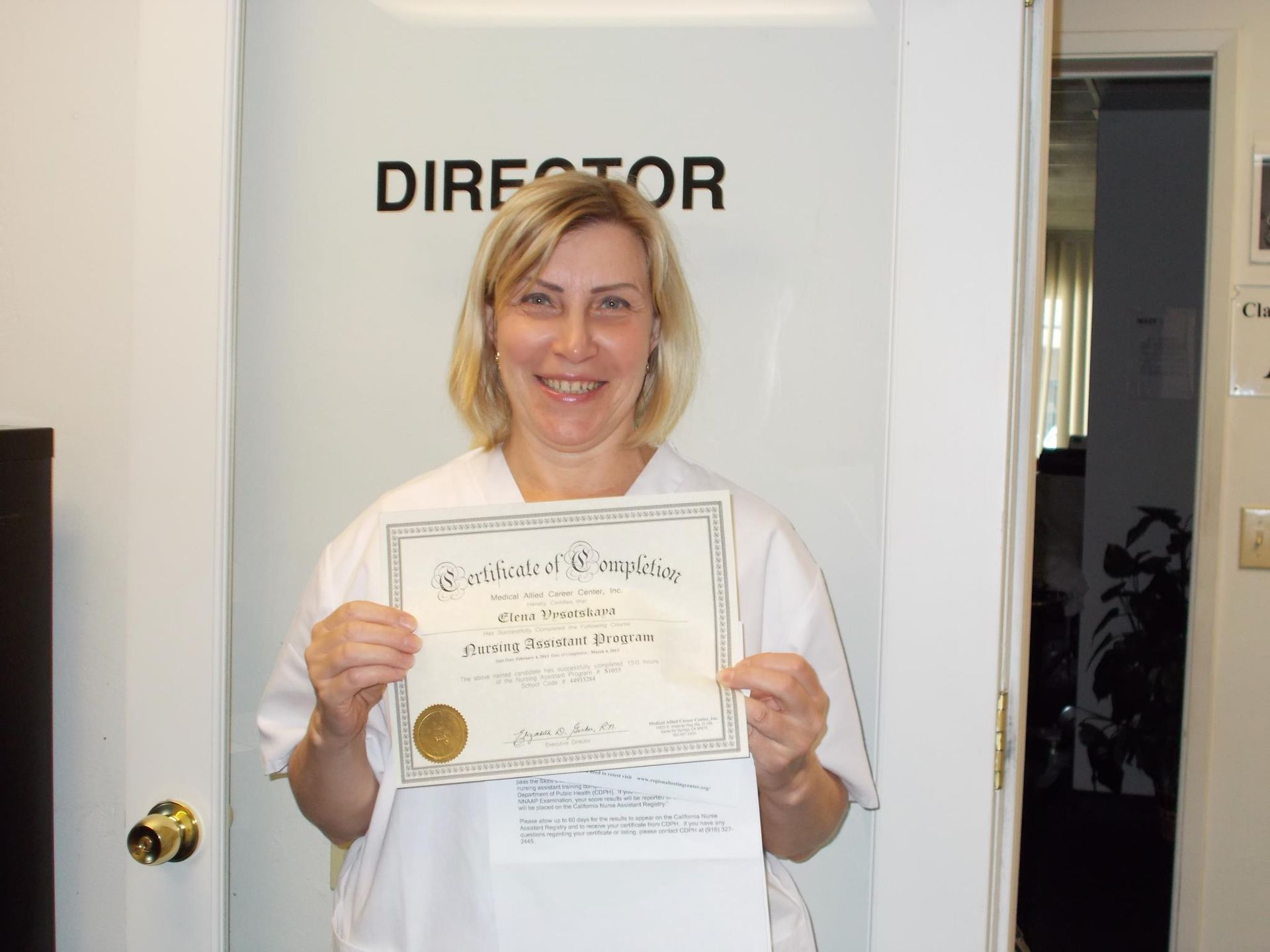 Woman in white uniform holds a certificate, smiling, in front of a door labeled