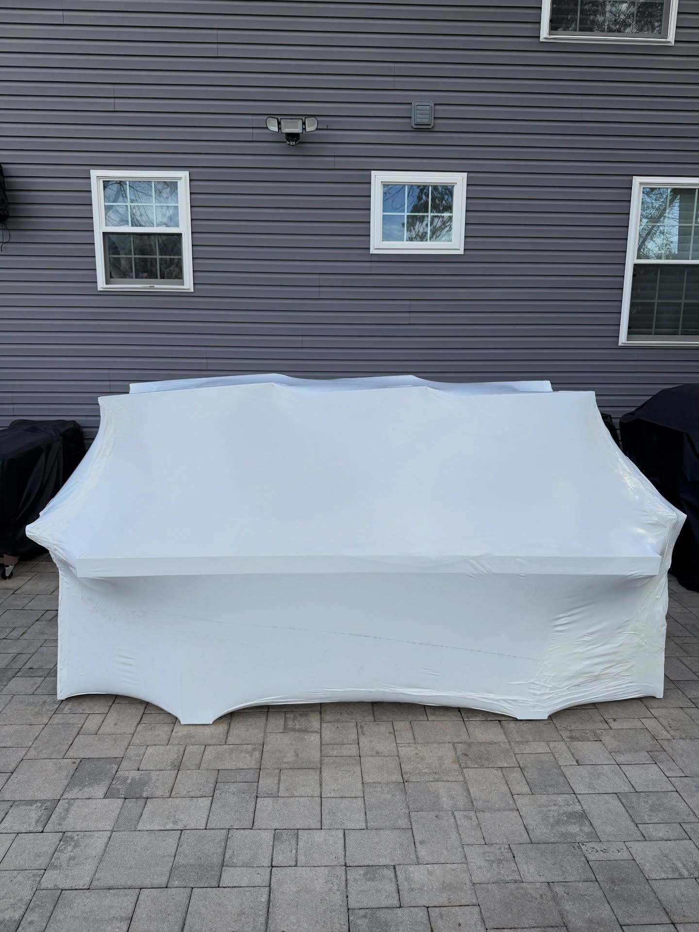 White table, covered in plastic wrap, sits on a brick patio in front of a gray building with windows.