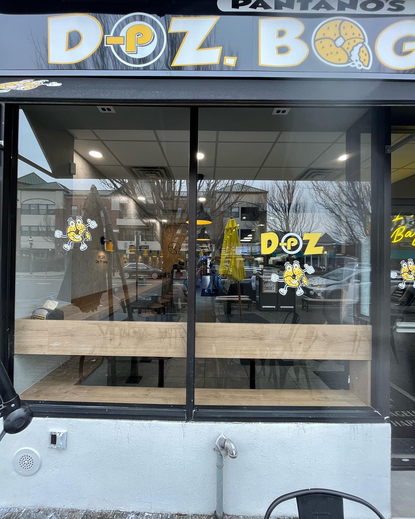 Doz. Bagels storefront with a large window, wooden benches, and bee-themed decorations; exterior view.