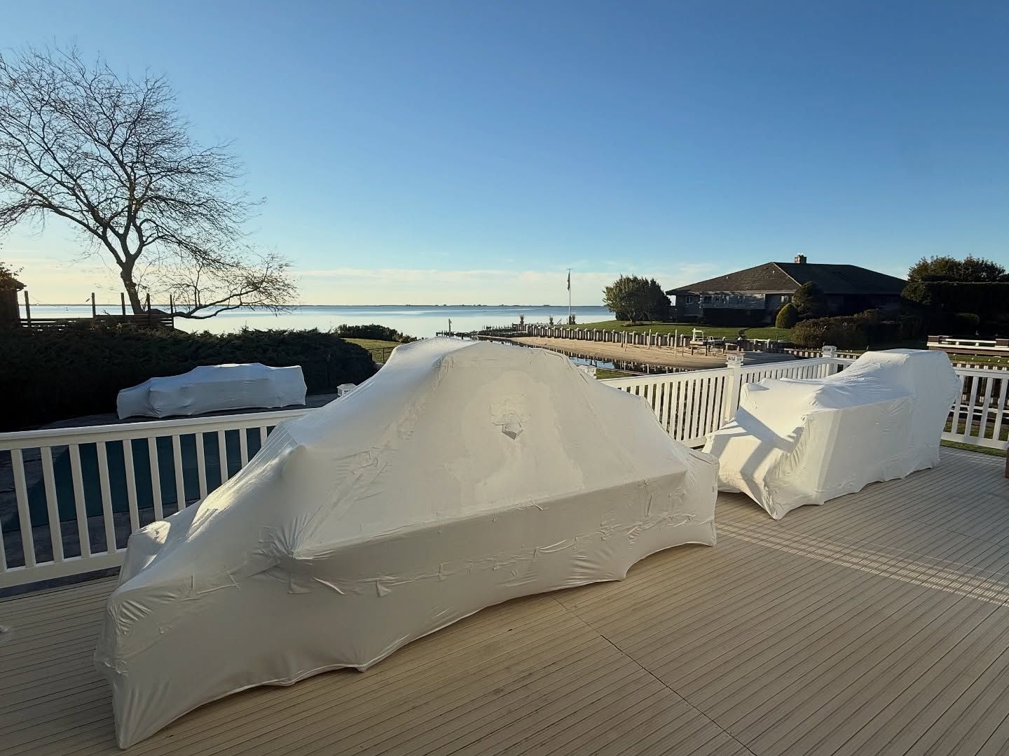 Outdoor furniture covered in white tarps on a wooden deck overlooking water on a sunny day.