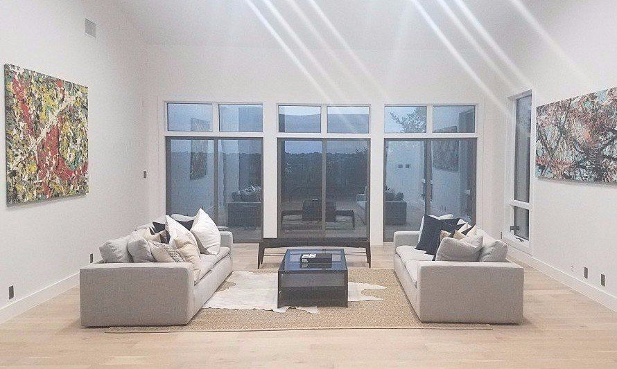 Bright modern living room with white sofas, coffee table, abstract art, and tall windows overlooking dusk outside