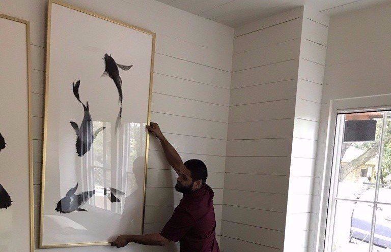 Person hanging framed fish art on a white paneled wall near a window.