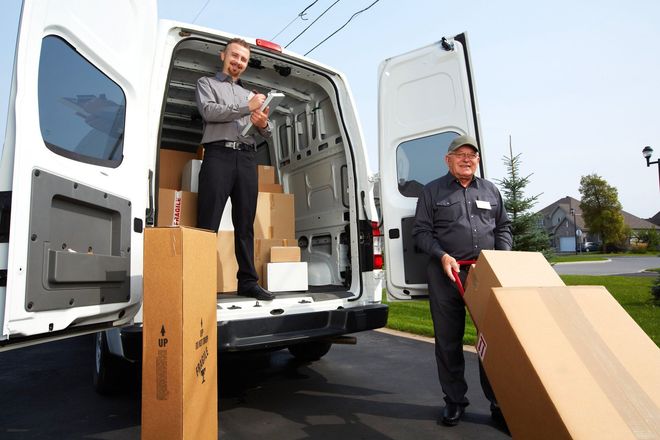 Two workers unloading large cardboard boxes from a white delivery van outdoors