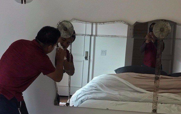 Two people inspect a bed in a bedroom, one holding a mirror near the headboard.