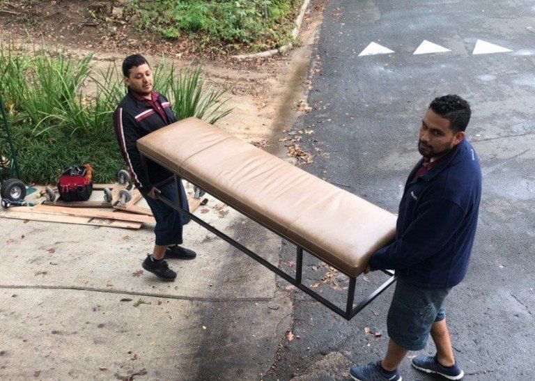 Two men carrying a long beige bench on a paved path outdoors
