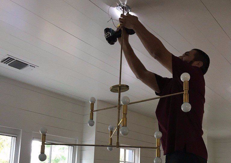 Man hanging a ceiling light fixture indoors using a ladder and reaching up with both hands