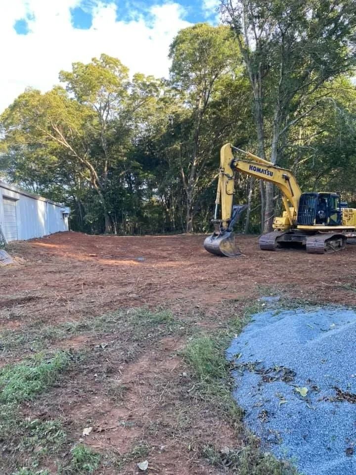 Yellow excavator on a cleared construction site with trees in the background. Gravel and dirt visible.