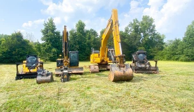Five yellow and black construction machines on grass field; trees in background.