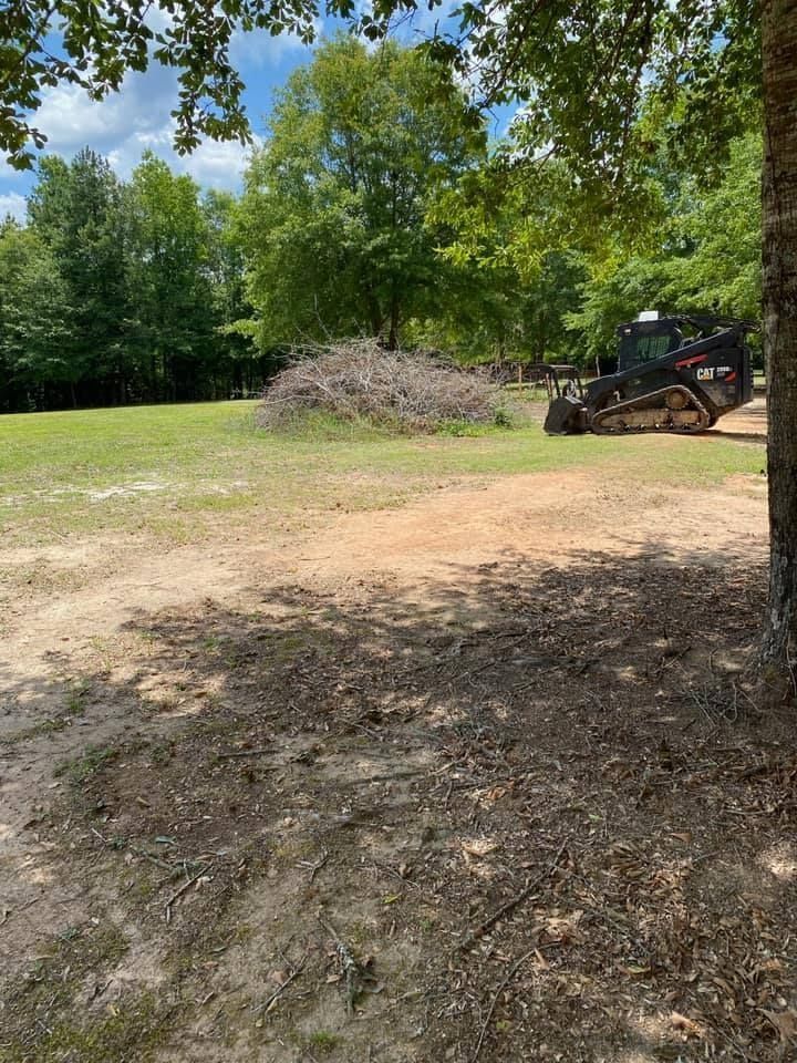 A small skid steer tractor near a pile of brush in a grassy field with trees.