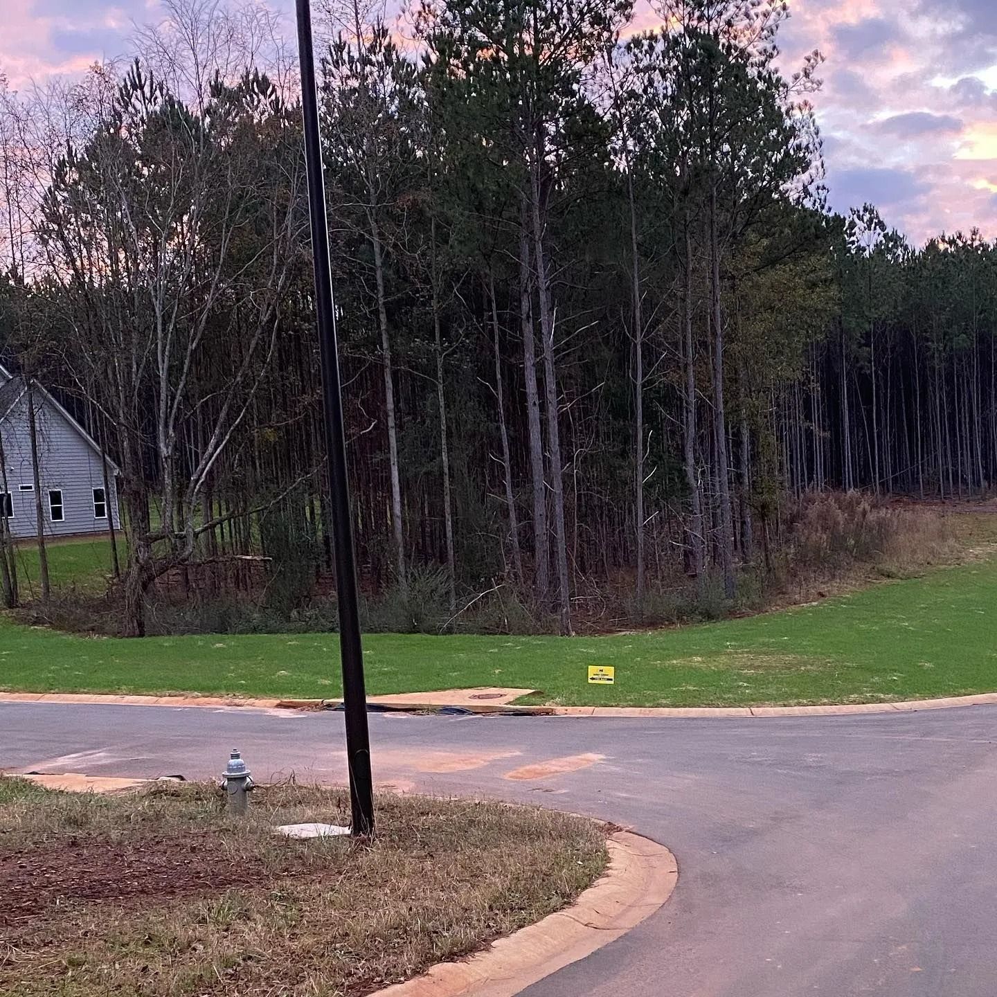 A paved road curves in front of a grassy area, trees, and a house under a colorful sky.