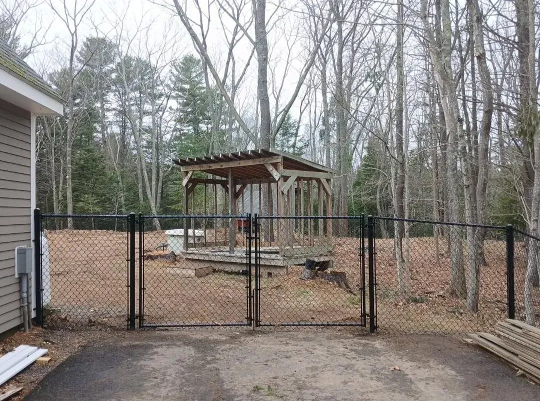 Backyard fenced area with a wooden pergola and picnic table, surrounded by trees and a house driveway.