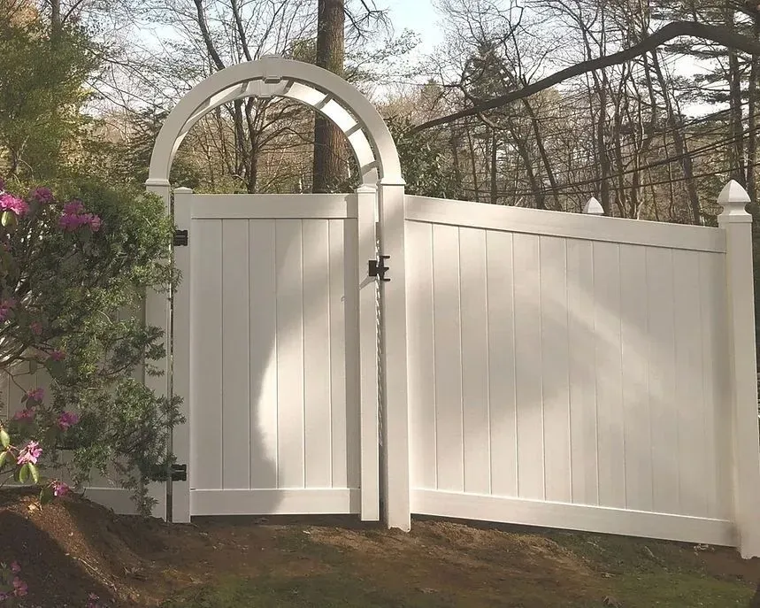 White arched wooden gate and fence beside blooming pink bushes in a wooded yard