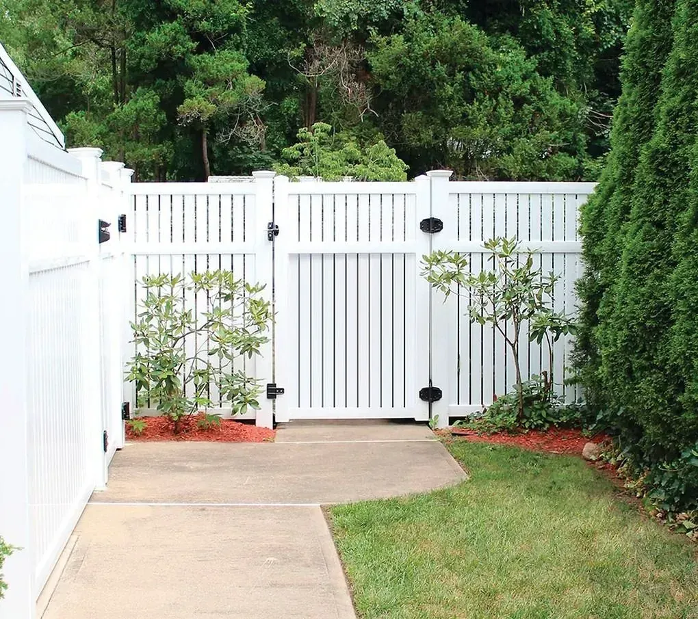 White backyard fence with gate beside a concrete path and green lawn, bordered by shrubs and trees