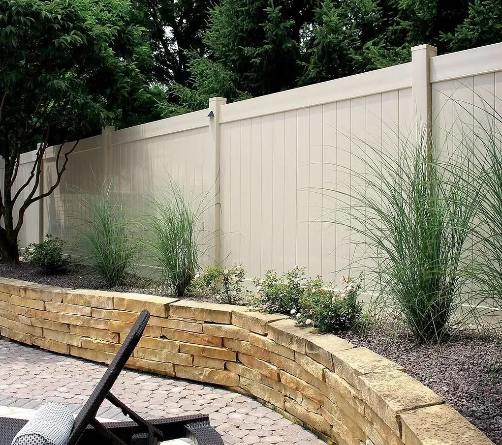 Curved stone retaining wall with sparse shrubs along a beige fence in a backyard patio area