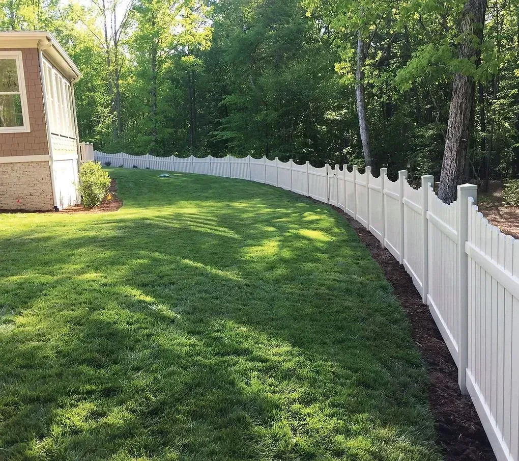 White vinyl fence curving along a green lawn beside a house and wooded yard