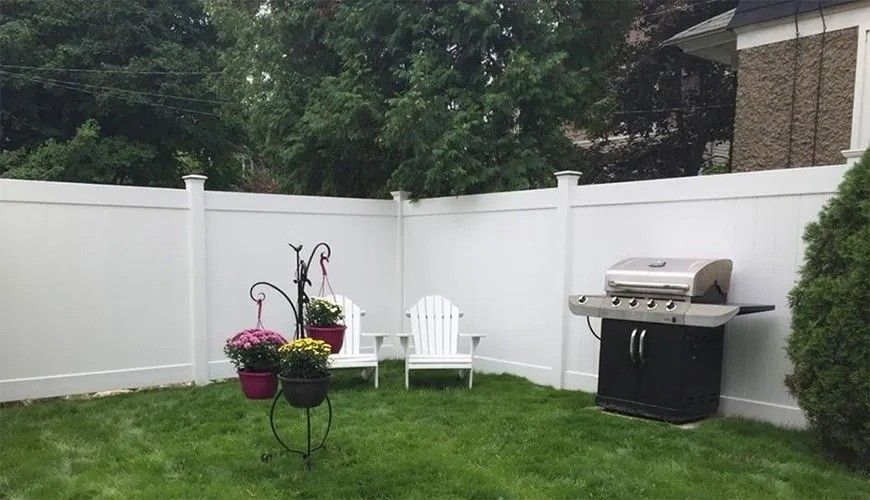 White fenced backyard with green lawn, patio chairs, hanging flowers, and a gas grill.
