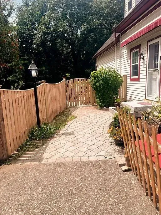 Curved brick walkway beside a house with wooden fences, a lamp post, and greenery in a sunny yard