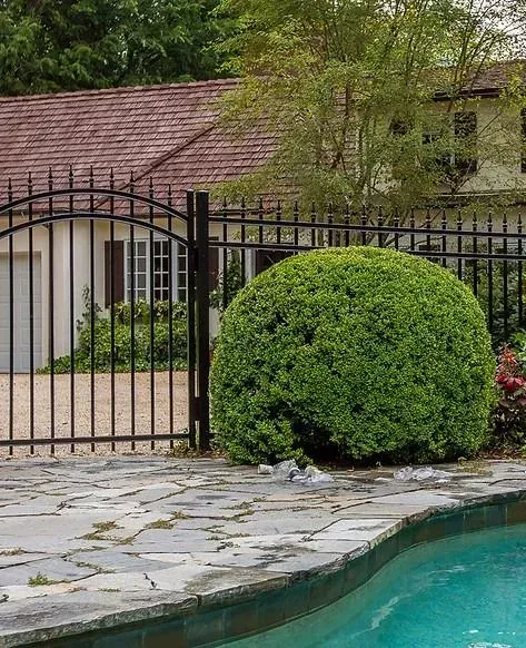 Backyard pool with stone deck, black fence, trimmed bush, and house in the background
