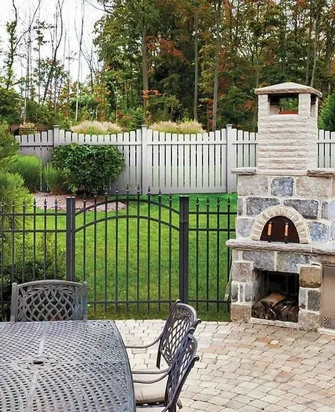 Backyard patio with metal table, black fence, white privacy fence, stone outdoor oven, and trees beyond