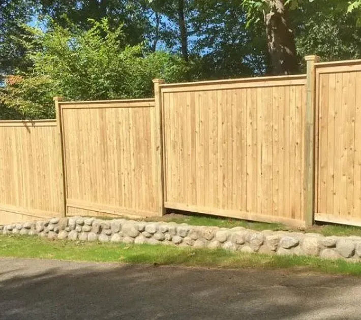 New wooden privacy fence with stone base along a paved road, backed by trees