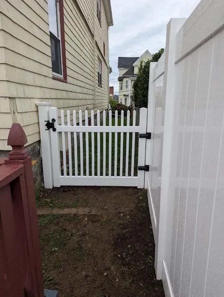 Narrow side yard with white picket gate, beige siding, and a tall white fence along a dirt path.