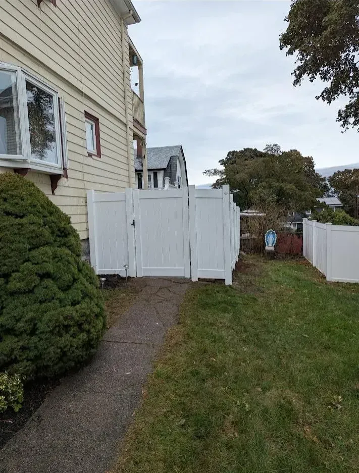 Narrow backyard path beside a beige house, leading to a white gate and fence under a cloudy sky.