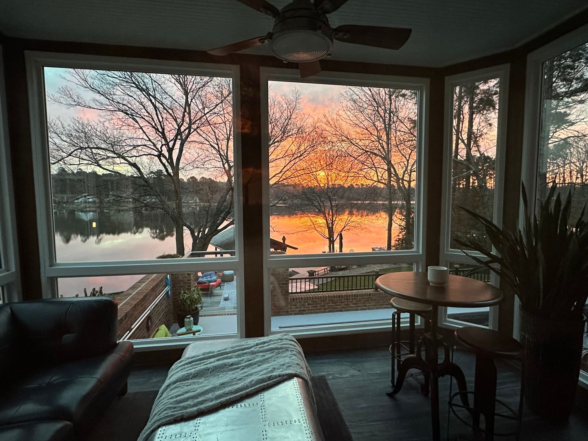 Sunroom with lake view at sunset; orange and pink sky, trees, table, couch.