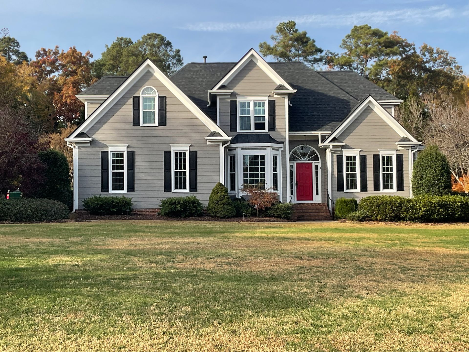 Two-story house with gray siding, black shutters, and a red front door, set on a green lawn with fall trees.
