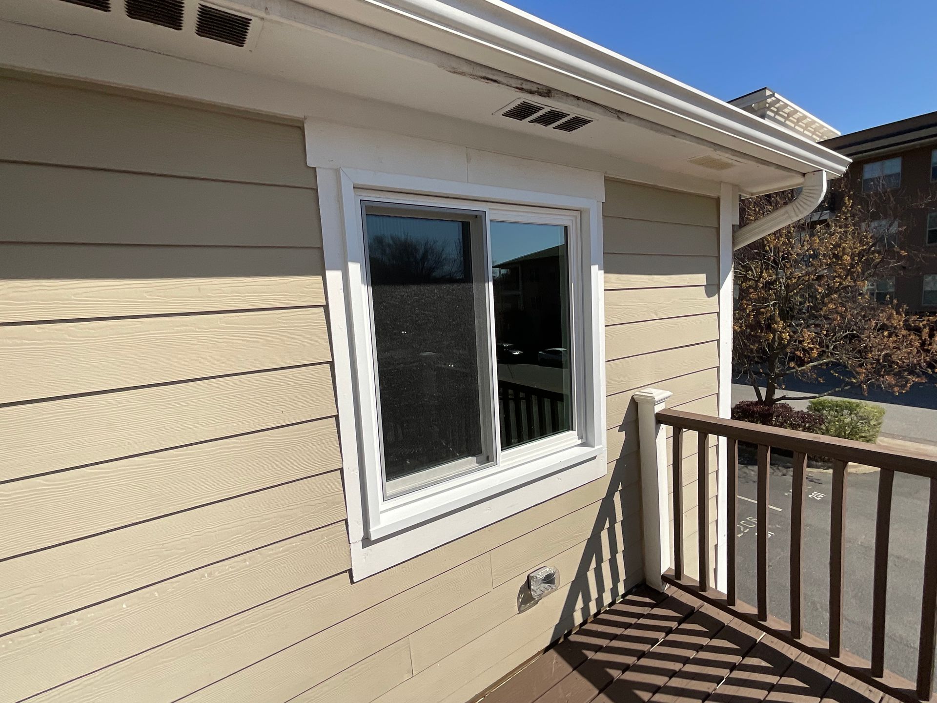 Tan siding with a white-framed window on a building's exterior, beside a brown deck railing.