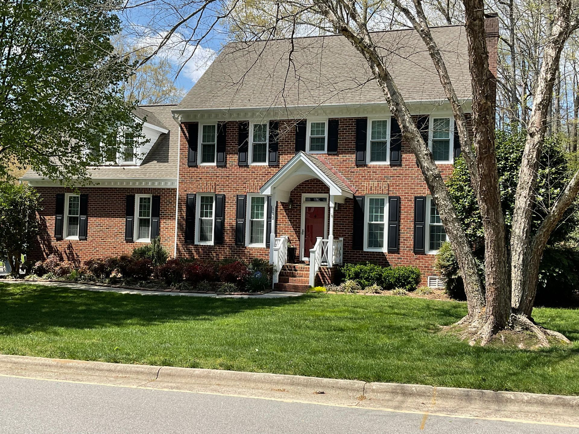 Brick house with black shutters, red door, green lawn, trees, and blue sky.
