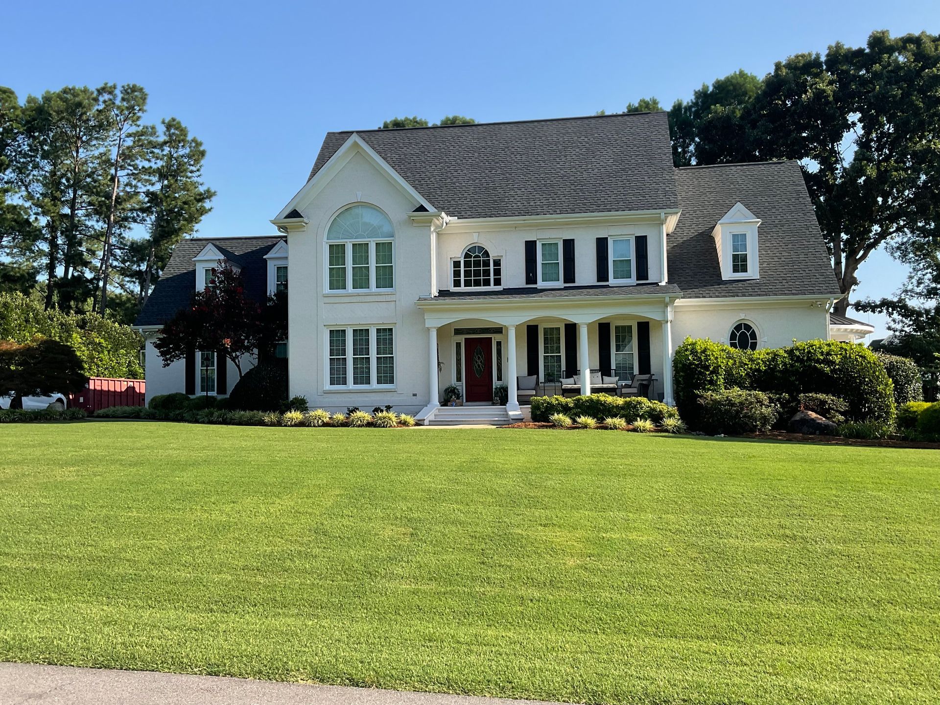White house with black shutters, columns, and a red door, on a grassy lawn under a blue sky.