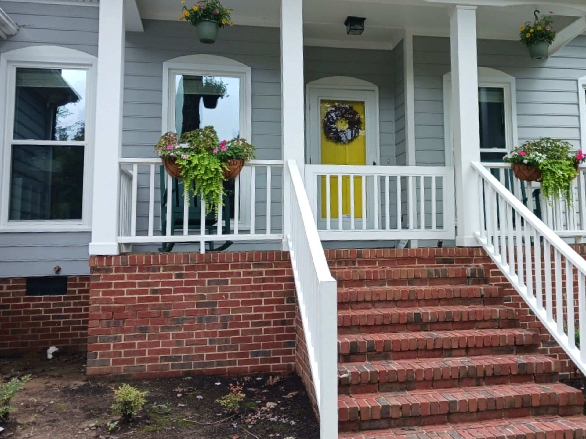 A house with a yellow door, white railing, and brick steps. Hanging flower baskets and a wreath decorate the porch.