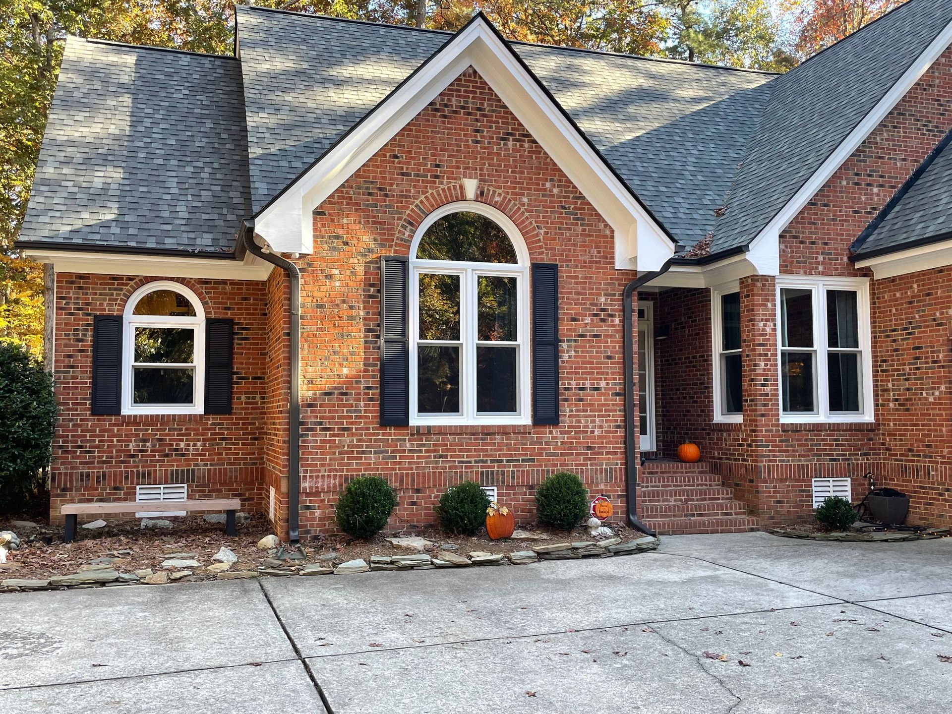 Brick house with white-framed windows, black shutters, and a dark gray roof. Pumpkins sit on the porch.