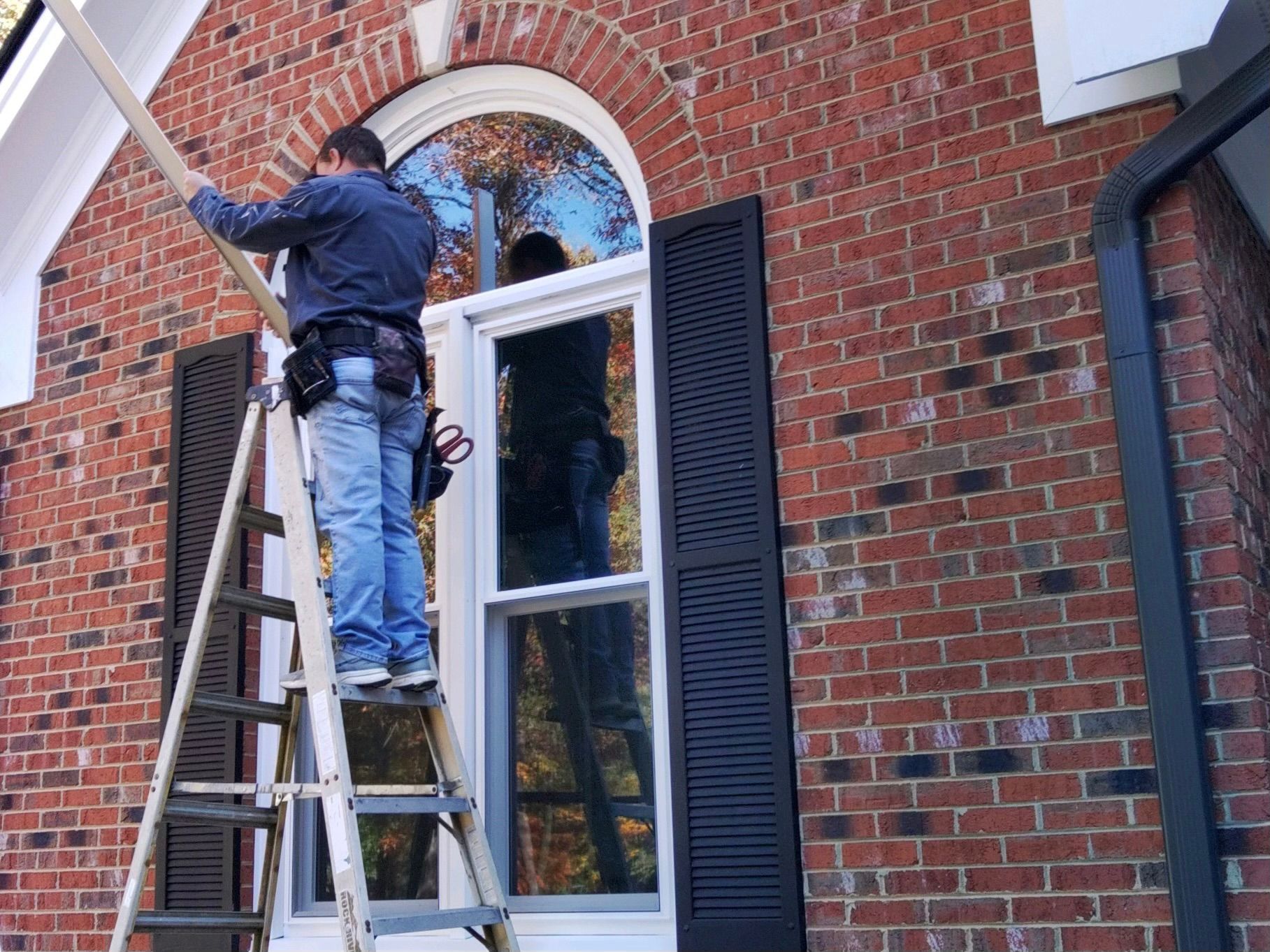 Man on ladder installing trim around a window on a brick building.