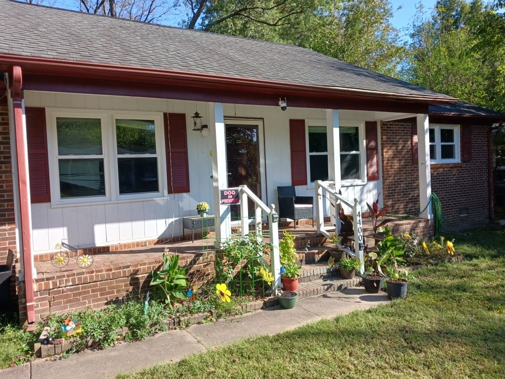 Brick bungalow with red shutters and trim, white porch, and small front yard with plants.