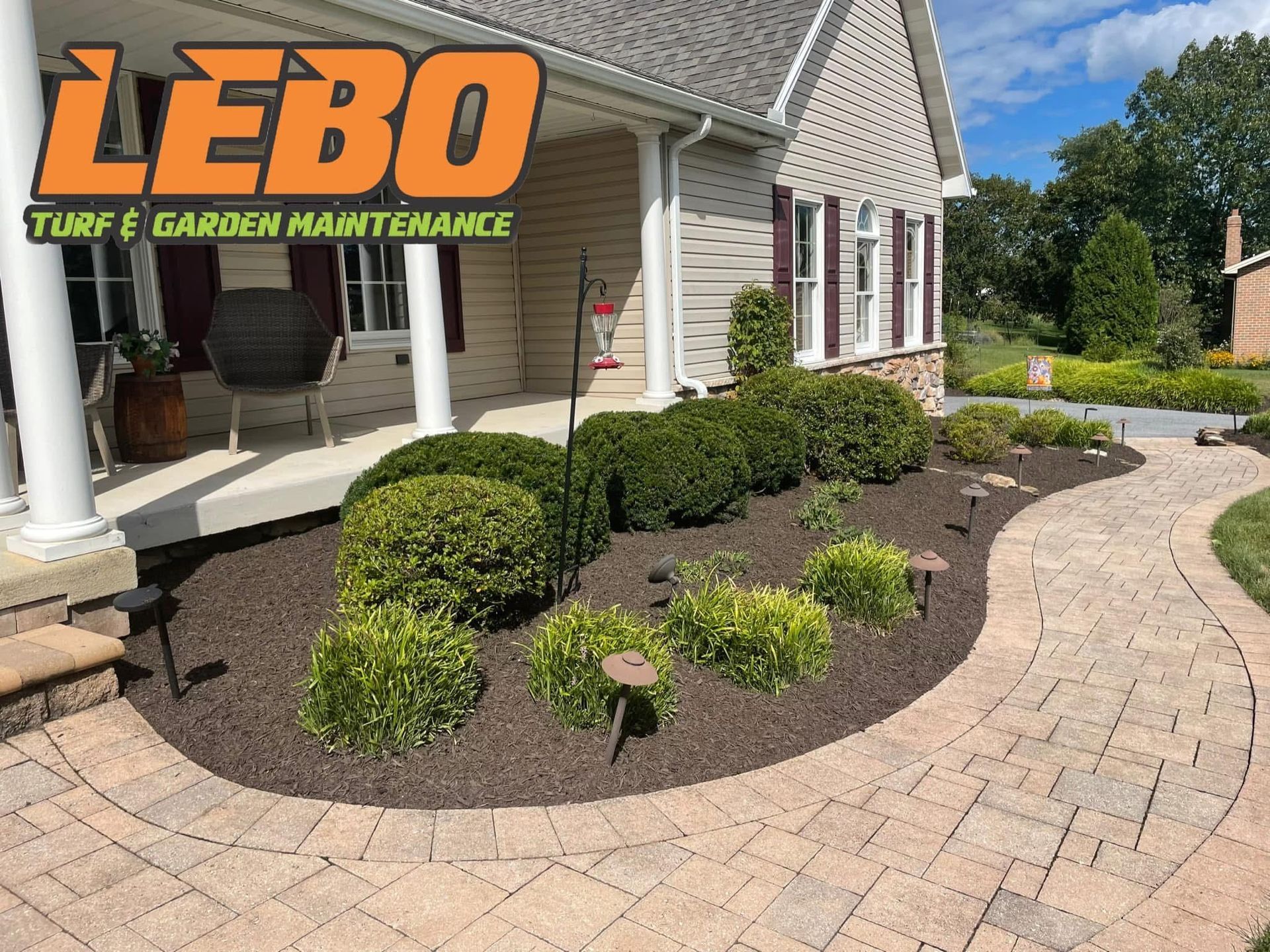 Front yard with mulch, bushes, and a brick path leading to the house; the logo 