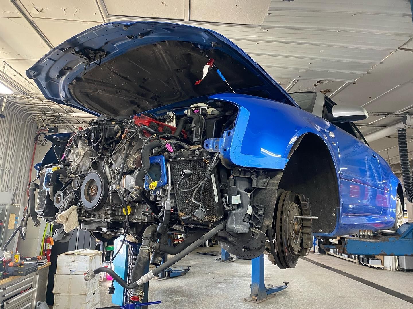 A blue car with the hood up is being worked on in a garage.