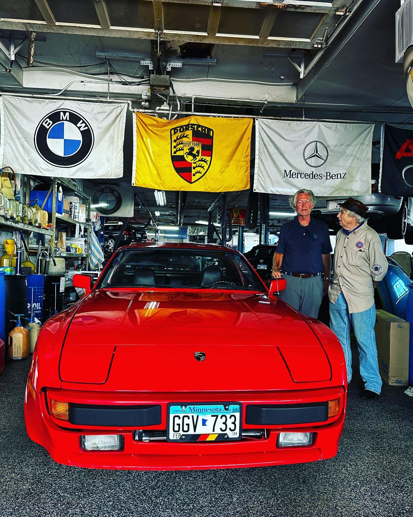 Two men are standing next to a red sports car in a garage.