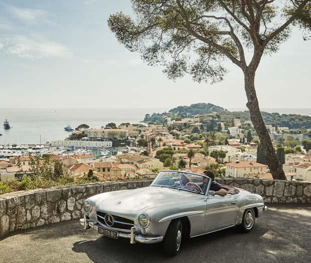 A man is driving a silver convertible car on a hill overlooking a city.