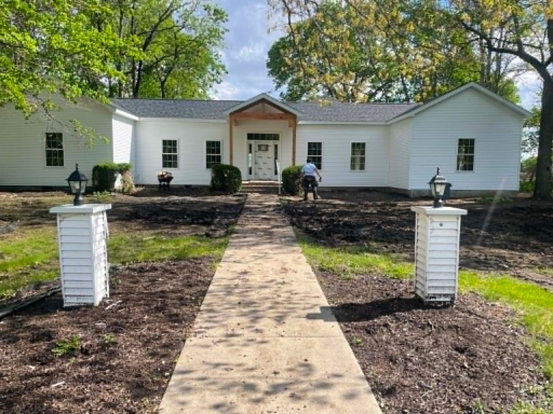 White house with a front walkway, flower beds, and two white brick pillars under trees.