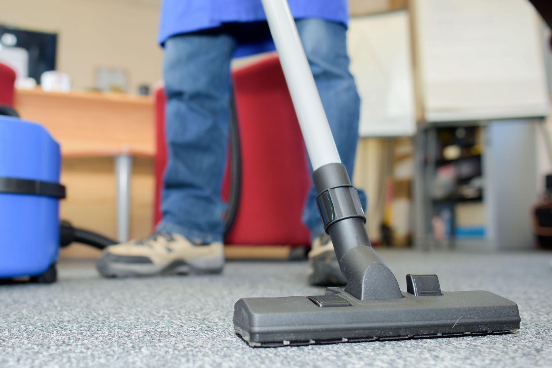 Person vacuuming carpet in an office; blue vacuum, red chair, desk.