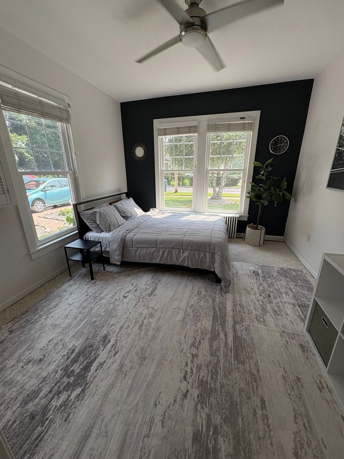 Bedroom with a bed against a black accent wall and a large window. Gray and white patterned rug and a ceiling fan.