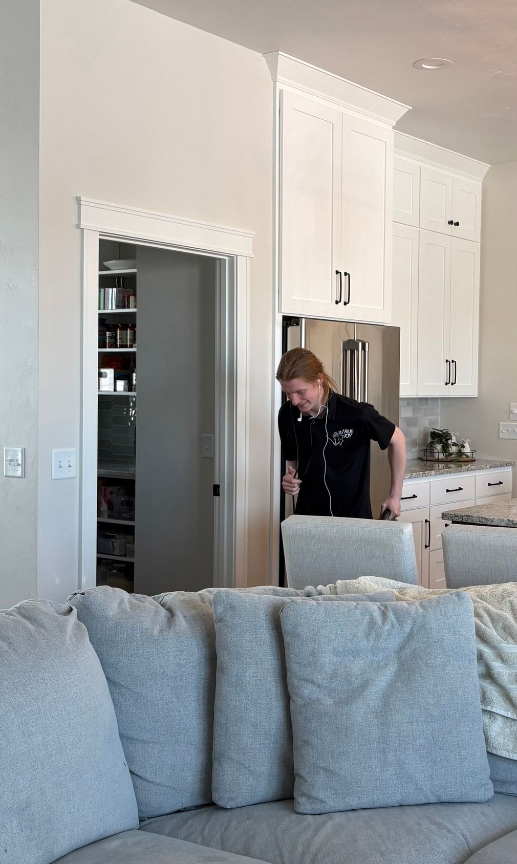 Person in black shirt, smiling, exiting a pantry into a kitchen. White cabinets, gray couch visible.