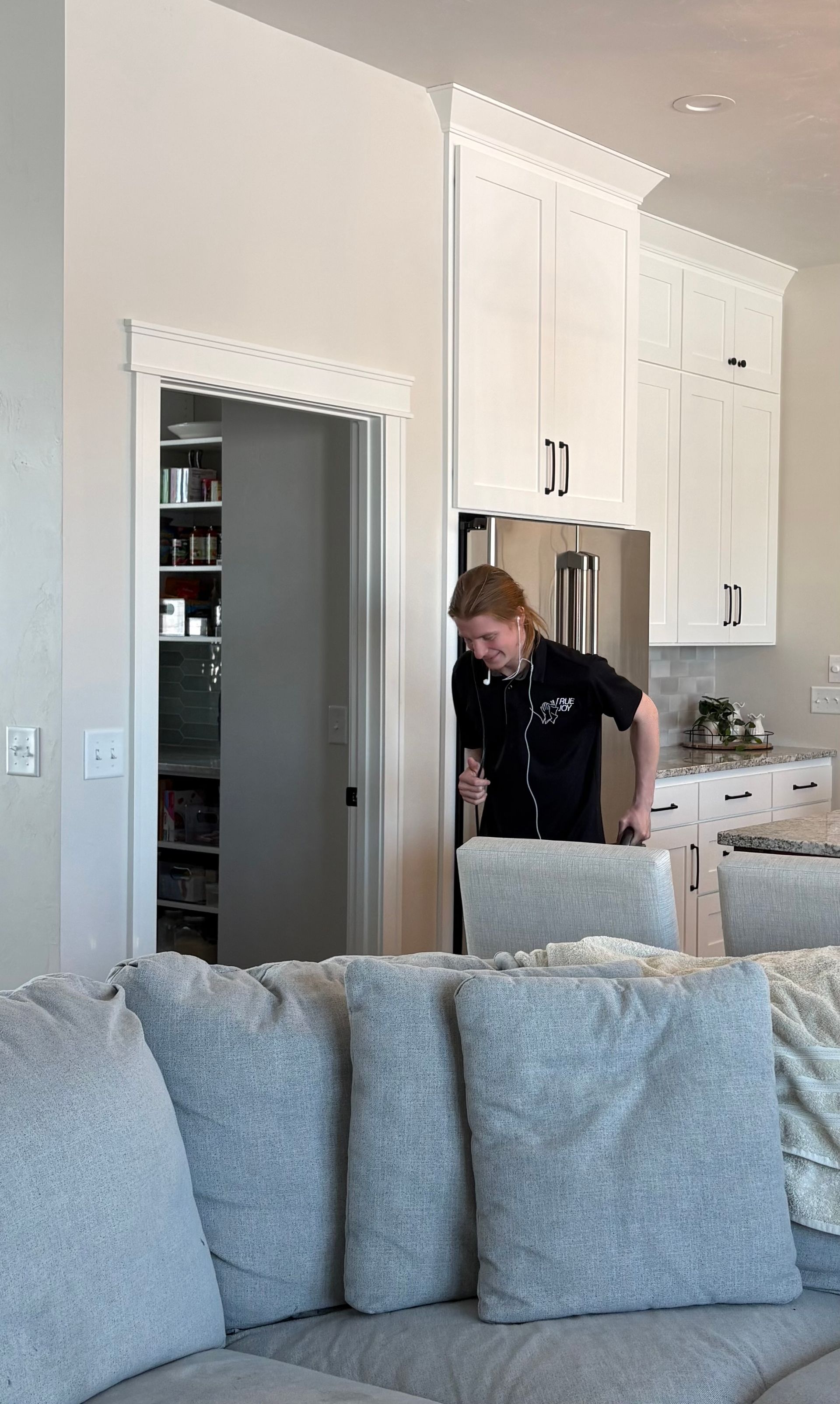 Person in black shirt, smiling, exiting a pantry into a kitchen. White cabinets, gray couch visible.