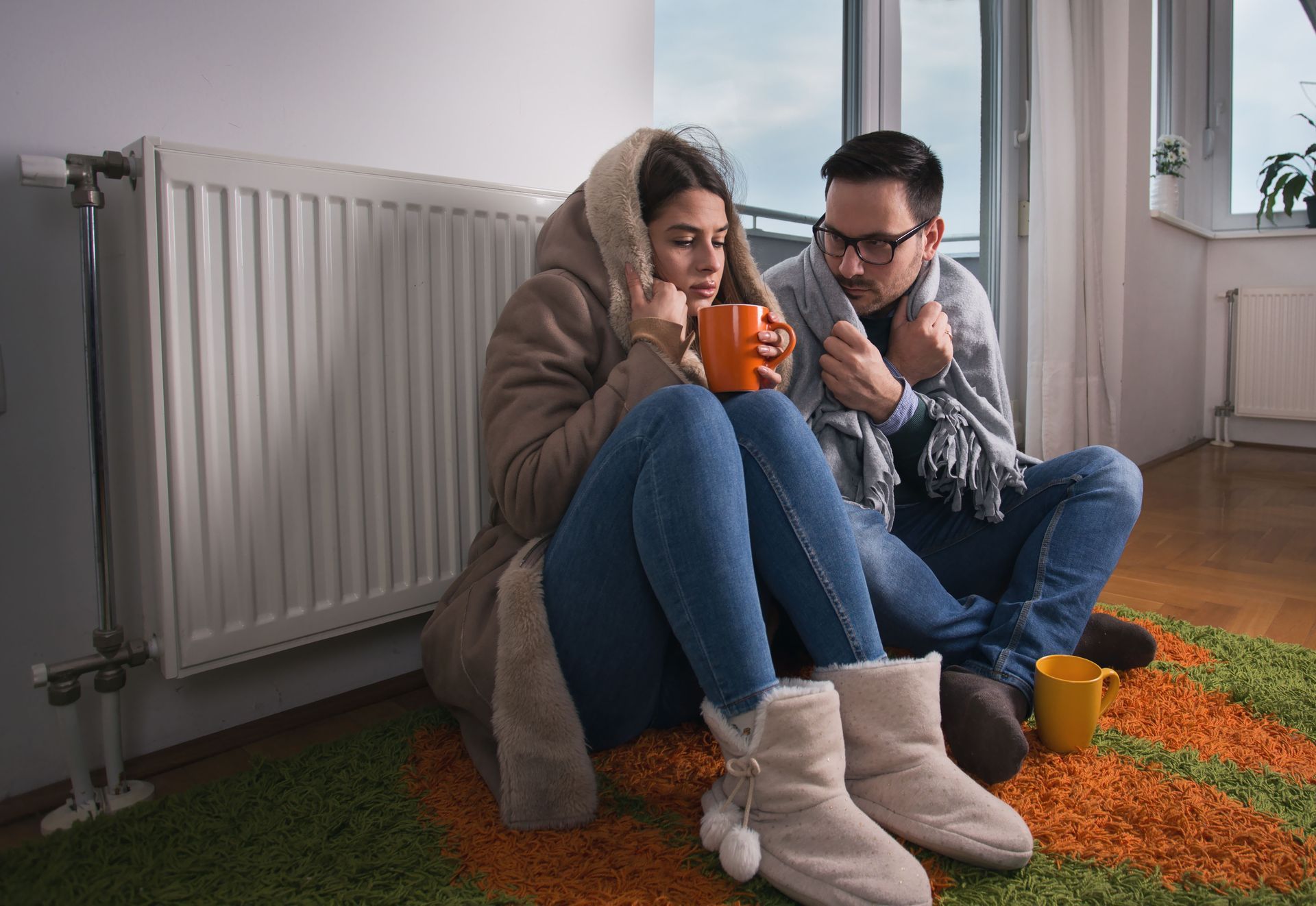 Couple huddled together near a radiator, wrapped in coats and blankets, holding mugs. Cold-looking.