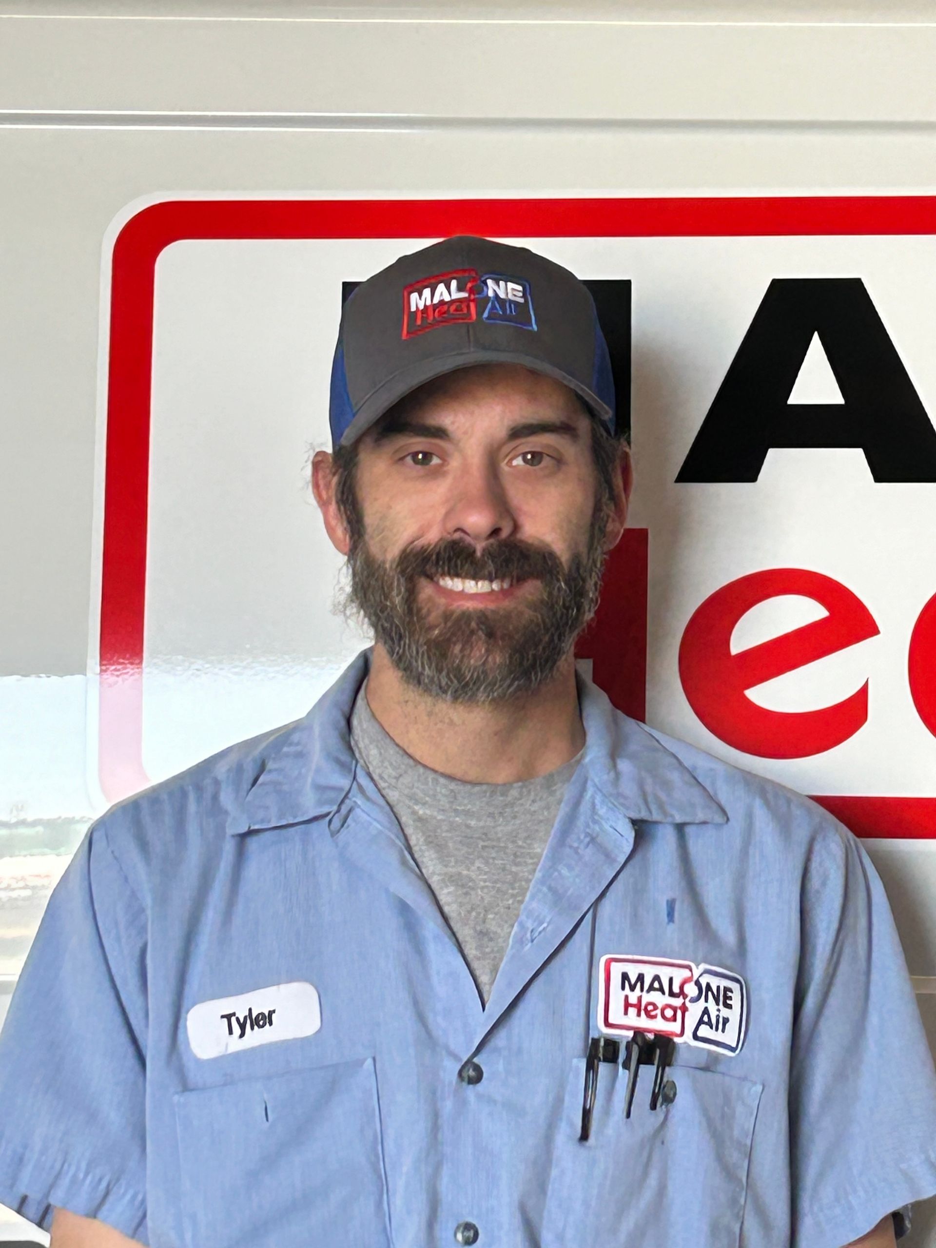 Man in a blue work shirt and cap, smiling in front of a sign.